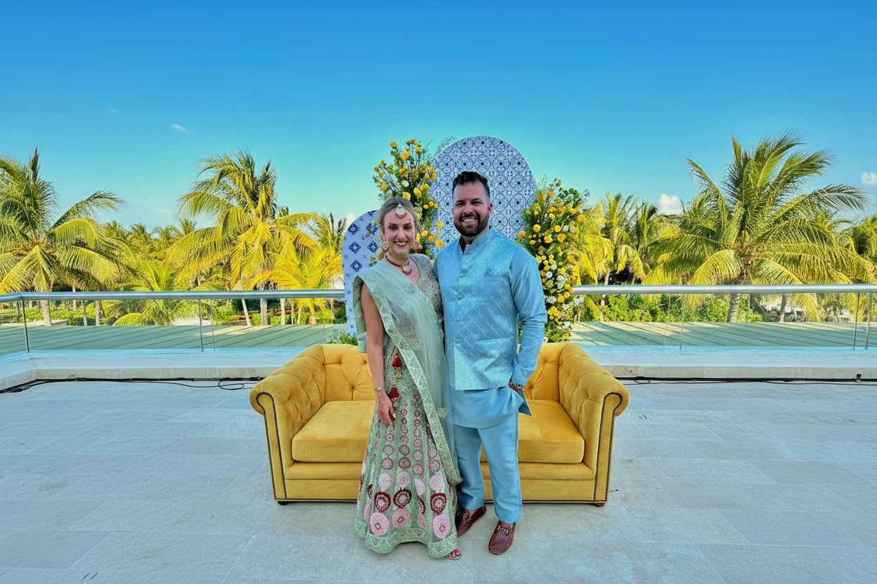 Man and woman in formal attire standing in front of decorative elements with palm trees in the background.